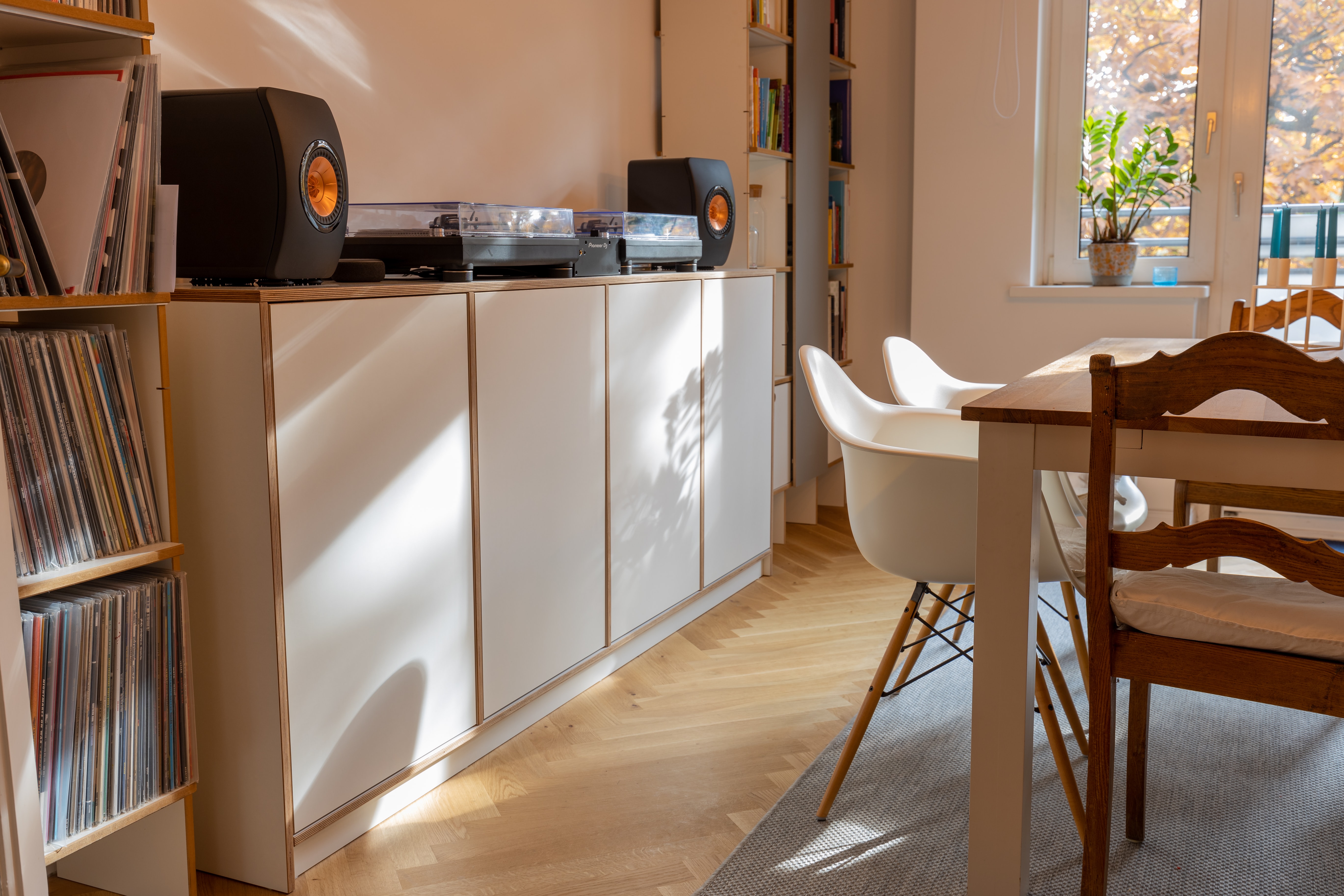 Brown and white cabinets , with cocoa brown seats, white chairs, light grey rug, small bookshelf with speakers and stereos, egg white window frame with sunlight coming through. 